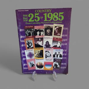 Könnte beinhalten: Ein Vintage-Musikbuch mit dem Titel "COUNTRY 25 OF 1985" von Columbia Pictures Publications. Das Cover ist lila mit grünem Text und zeigt Bilder verschiedener Country-Musikkünstler und Songtitel. Das Buch wird auf einem transparenten Ständer präsentiert.