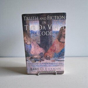 May include: A hardcover book titled "Truth and Fiction in The Da Vinci Code" by Bart D. Ehrman. The book cover features a painting with figures and text. The book is displayed on a small metal stand.