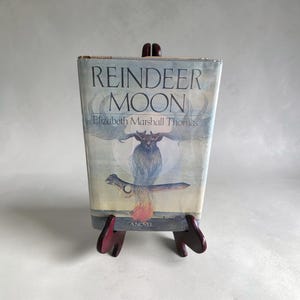 Puede incluir: Un libro de tapa dura titulado Reindeer Moon de Elizabeth Marshall Thomas, con una sobrecubierta que presenta una ilustración en azul pálido y blanco de una cabeza de reno, astas grandes y una luna brillante, sobre un soporte de madera marrón.