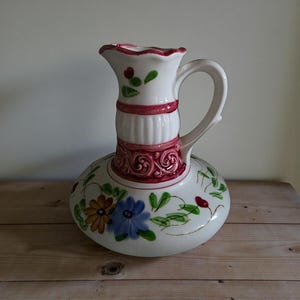May include: A white ceramic pitcher with a bulbous base and a handle. The pitcher is decorated with floral designs in blue, orange, and green. It has a red band around the middle and a ruffled rim. The pitcher is approximately 12 inches tall.