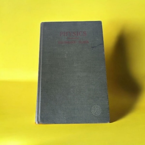 Puede incluir: Un libro antiguo titulado "PHYSICS" en letras rojas en la portada. El libro tiene una cubierta gris oscura y está sobre un fondo amarillo brillante. El lomo del libro es visible y un pequeño emblema está en la esquina inferior derecha.