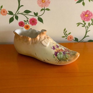 Puede incluir: Jarrón de porcelana con forma de zapato, de color crema, con un diseño floral que presenta flores moradas y verdes. El borde y los detalles tienen un acabado dorado. El zapato está sobre una superficie de madera.