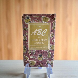 May include: A vintage cookbook titled "The ABC of Herb & Spice Cookery" by Peter Pauper Press. The cover is a maroon color with illustrations of cooking tools and spices. The title is in a gold color, and the book is displayed on a clear plastic stand.