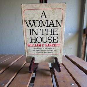 May include: A hardcover book titled "A Woman in the House" by William E. Barrett, displayed on a wooden stand. The book has a cream-colored cover with red and black text. The stand is made of dark wood.