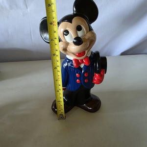 K&ouml;nnte beinhalten: Eine Keramikfigur von Mickey Mouse, etwa 25 cm hoch, gekleidet in einem blauen Anzug mit roten Akzenten. Die Figur h&auml;lt einen schwarzen Zylinderhut und hat einen l&auml;chelnden Ausdruck. Die Figur steht auf einer wei&szlig;en Oberfl&auml;che.
