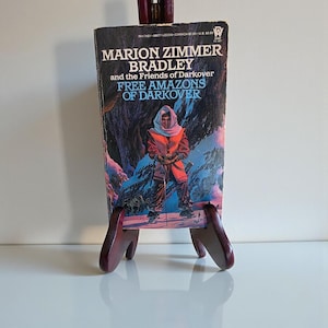 May include: A paperback book titled "Marion Zimmer Bradley and the Friends of Darkover: Free Amazons of Darkover" is displayed on a wooden easel. The book cover features a person in red and orange clothing standing in a snowy landscape.