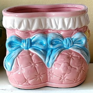 Op de afbeelding: Een roze keramische plantenpot in de vorm van babyschoentjes met blauwe strikken en witte rand.