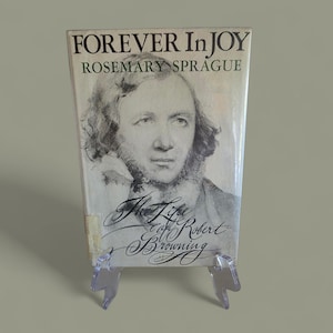 May include: A vintage book titled "Forever In Joy" by Rosemary Sprague. The cover features a portrait of a man and the title "The Life of Robert Browning". The book is displayed on a clear plastic stand.
