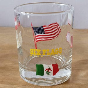 Könnte beinhalten: Ein durchsichtiger Glasbecher mit einem Six Flags Logo und verschiedenen Flaggen, die auf die Seite gedruckt sind. Zu den Flaggen gehören die amerikanische Flagge, die mexikanische Flagge und eine weiße Flagge mit einem roten Streifen.