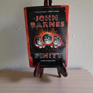 May include: A book titled "FINITY" by John Barnes, displayed on a wooden stand. The cover features a dark background with planetary graphics and the author's name in bold letters. The book is a "novel of many worlds."