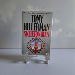 Pode incluir: Um livro de capa dura intitulado "SKELETON MAN" de Tony Hillerman. O livro tem uma capa prateada com texto preto e vermelho. Um design geométrico colorido é apresentado na parte inferior da capa. O livro é exibido em um suporte de plástico transparente.