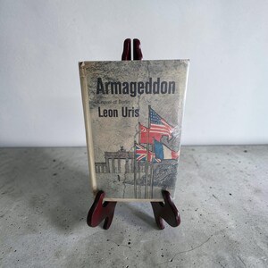 Puede incluir: Una copia vintage de "Armageddon: A Novel of Berlin" de Leon Uris, exhibida en un pequeño caballete de madera rojo oscuro. La portada del libro presenta banderas y una representación de la Puerta de Brandeburgo. El libro se sujeta con un soporte de madera y una goma elástica.