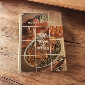 May include: A vintage cookbook titled "Good Housekeeping Menus for a Whole Year of Dinners". The cover features a colorful photo of a dinner plate with a variety of foods, including meat, vegetables, and a side of peas.