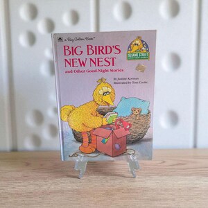 May include: A Big Golden Book titled "Big Bird's New Nest and Other Good-Night Stories." The cover features an illustration of Big Bird, a yellow Sesame Street character, with a nest and a red box. The book is propped up on a clear stand.