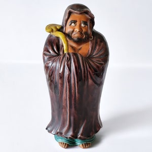 May include: A ceramic figurine of a monk with a brown robe and a yellow staff. The monk has a serious expression on his face and is holding his hands together in prayer.