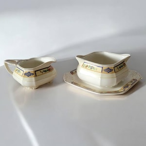 May include: A cream-coloured ceramic creamer and gravy boat set. The pieces feature a yellow band with floral and geometric designs. The creamer has a handle and sits on three small feet. The gravy boat rests on a matching rectangular plate.