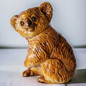 Könnte beinhalten: Eine braune Keramikfigur eines Koalas mit einem strukturierten Fellmuster. Der Koala sitzt mit dem Kopf zur Seite geneigt und blickt nach vorne.
