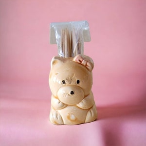 May include: A ceramic bear toothpick holder with a pink bow on its head. The holder is filled with wooden toothpicks.