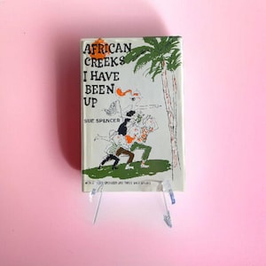 Può includere: Un libro vintage intitolato "African Creeks I Have Been Up" di Sue Spencer. La copertina presenta un'illustrazione a cartoni animati di persone e una palma. Il libro è esposto su un supporto in acrilico trasparente su sfondo rosa.
