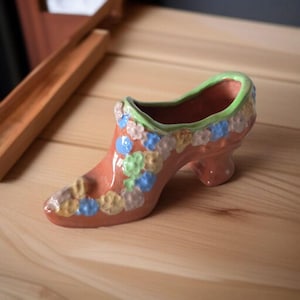 May include: A ceramic shoe-shaped planter in a peach color, adorned with colorful floral accents. The interior rim is green, and the shoe rests on a wooden surface. A decorative item for home decor.