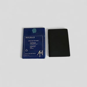 Può includere: Una scatola blu e un libro nero sono esposti su una superficie bianca. La scatola, etichettata "HOLMAN ROYAL RUBY TEXT BIBLE", presenta testo bianco e un logo. Il libro è chiuso e ha una copertina nera liscia. La scatola è aperta.