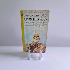 May include: A vintage paperback book titled "UPON THIS ROCK" by Frank G. Slaughter. The cover features an illustration of a man with a white beard and a yellow border. The book is displayed on a clear acrylic stand.