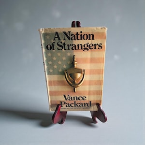 May include: A vintage book titled "A Nation of Strangers" with an American flag design and a brass door knocker. The book is displayed on a small, dark red wooden stand.