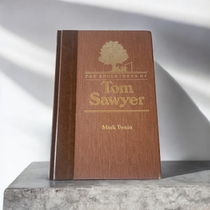 May include: A brown hardcover book with a gold title, "The Adventures of Tom Sawyer" by Mark Twain. The cover features a stylized tree with a small house in the background.