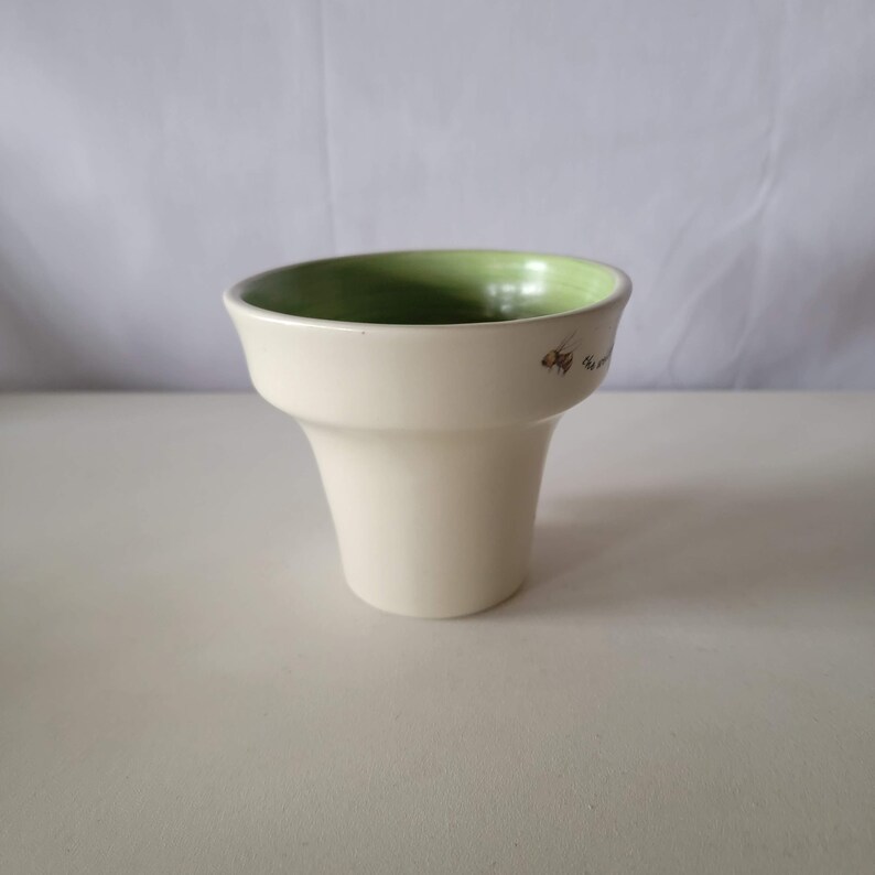 May include: A cream-colored ceramic planter with a flared shape. The interior is painted a light green. Small brown text and a graphic of a bee are visible on the side of the planter. The planter is on a white surface.