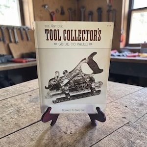 May include: A softcover book titled The Antique Tool Collector's Guide to Value by Ronald S. Barlow. The off-white cover features a detailed black and white illustration of an ornate metal hand plane with a dark handle, centered on the front.