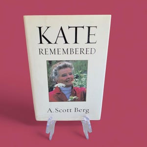 May include: A hardcover book titled "KATE REMEMBERED" by A. Scott Berg. The book cover features a portrait of a woman in a red jacket. The book is displayed on a clear plastic stand against a pink background.