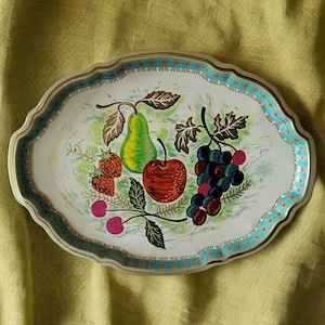 May include: A white oval metal tray with a blue and gold trim. The tray features a painted design of fruit including a pear, strawberries, cherries, and grapes.