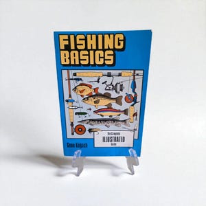 May include: A blue book titled "FISHING BASICS" with illustrations of fish and fishing gear. The book is subtitled "The Complete Illustrated Guide" and is displayed on a clear plastic stand.