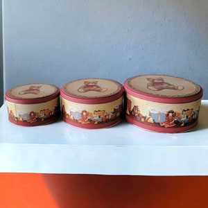 May include: Three round, red and cream colored metal storage tins with a teddy bear and toy train design. The tins are stacked on top of each other.