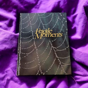 May include: A black book with a spiderweb design and the words "Fragile Moments" in gold lettering.