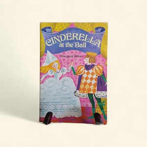 May include: A vintage children's book titled "Cinderella at the Ball" by Margaret Hillert. The cover features an illustration of Cinderella in a white gown and a prince in a checkered orange and purple outfit. The book is propped up by a black stand.
