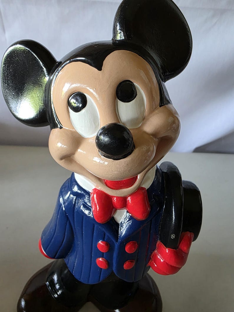 K&ouml;nnte beinhalten: Keramikfigur von Mickey Mouse, mit einem schwarzen Zylinder, einem blauen Anzug mit roten Kn&ouml;pfen und einer roten Fliege. Die Figur hat ein beiges Gesicht, schwarze Ohren und eine schwarze Nase. Die Figur h&auml;lt einen schwarzen Zylinder.