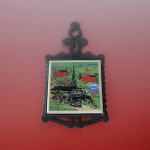 Puede incluir: Trípode de hierro fundido negro con una baldosa cerámica cuadrada que presenta una ilustración colorida de una iglesia, dos casas rojas y un paisaje verde.