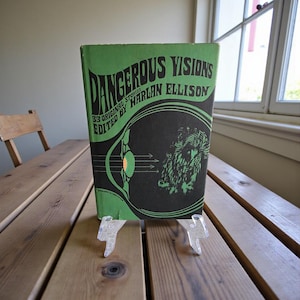 Puede incluir: Un libro antiguo de tapa dura titulado "Dangerous Visions" de Harlan Ellison. El libro tiene una cubierta verde con letras negras y una ilustración gráfica. Está apoyado en un soporte de plástico transparente sobre una mesa de madera.