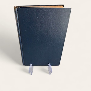 Pode incluir: Um livro azul marinho com uma capa desgastada e texturizada. A borda superior do livro é castanho claro. O livro é apoiado por dois suportes de plástico transparente. O livro está ligeiramente inclinado.