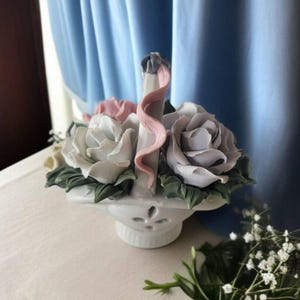 May include: A white porcelain flower arrangement in the shape of a basket with three roses. The roses are white, pink, and lavender. The basket has a decorative handle with a pink ribbon.
