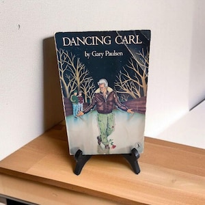 May include: A vintage paperback book titled "DANCING CARL" by Gary Paulsen. The cover art depicts a man in a brown jacket and green trousers, with a rose at his feet, set against a winter scene with leafless trees and two figures in the background.