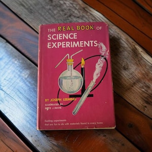 May include: A red book titled "The Real Book of Science Experiments" by Joseph Leeming, illustrated by Ruth T. Davis. The cover features a science experiment with a glass flask, a tube, and a lit candle.