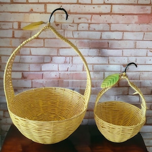 Könnte beinhalten: Zwei gelbe Hängekörbe aus Weidengeflecht in Form von Birnen. Der größere Korb hat einen schwarzen Metallgriff und der kleinere Korb hat einen schwarzen Metallgriff mit einem grünen Blatt.