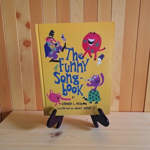 Op de afbeelding: Een vintage kinderboek getiteld "The Funny Song-book" met illustraties. De omslag is geel met kleurrijke cartoonfiguren en tekst. Het boek wordt gepresenteerd op een donkerbruine houten standaard tegen een lichtbruine houten achtergrond.