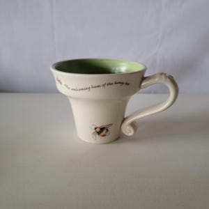 May include: Cream-colored ceramic mug with a flared shape and a curved handle. The interior is green, and the mug features a bee illustration and the text "the welcoming hum of the honey bee."