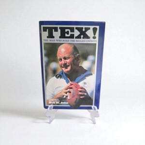 May include: A book titled "TEX! The Man Who Built the Dallas Cowboys" featuring a portrait of a man holding a football. The book has a blue border and is displayed on a clear plastic stand.