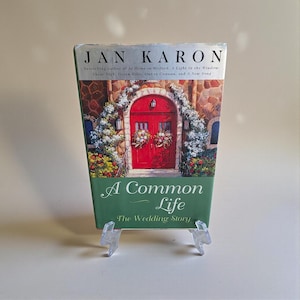 May include: A hardcover book titled "A Common Life: The Wedding Story" by Jan Karon. The cover features a red door with floral arrangements, set against a stone building. The book is displayed on a clear plastic stand.