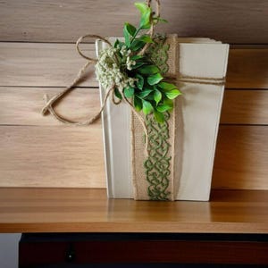 May include: A cream-colored, rectangular box decorated with a green burlap ribbon and twine. The box is adorned with faux greenery and small white flowers, creating a rustic, natural aesthetic. The box is displayed on a wooden shelf.
