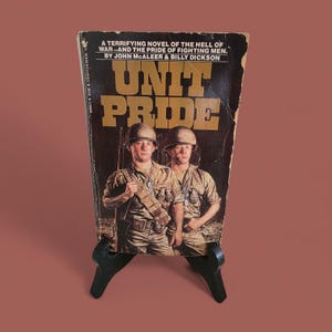 Könnte beinhalten: Vintage Taschenbuch mit dem Titel "Unit Pride" von John McAleer und Billy Dickson. Das Cover zeigt zwei Soldaten in Uniform und Helmen. Das Buch wird auf einem schwarzen Ständer vor braunem Hintergrund präsentiert.
